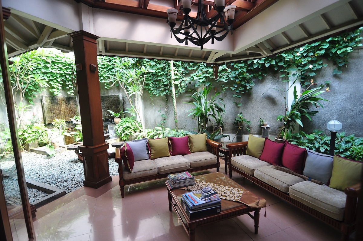 A serene living area features a comfortable seating arrangement with cushions in various colors. Lush greenery and a decorative water feature are visible through the open space, creating a calm atmosphere. Natural light filters in, highlighting the stylish wooden accents and serene surroundings.