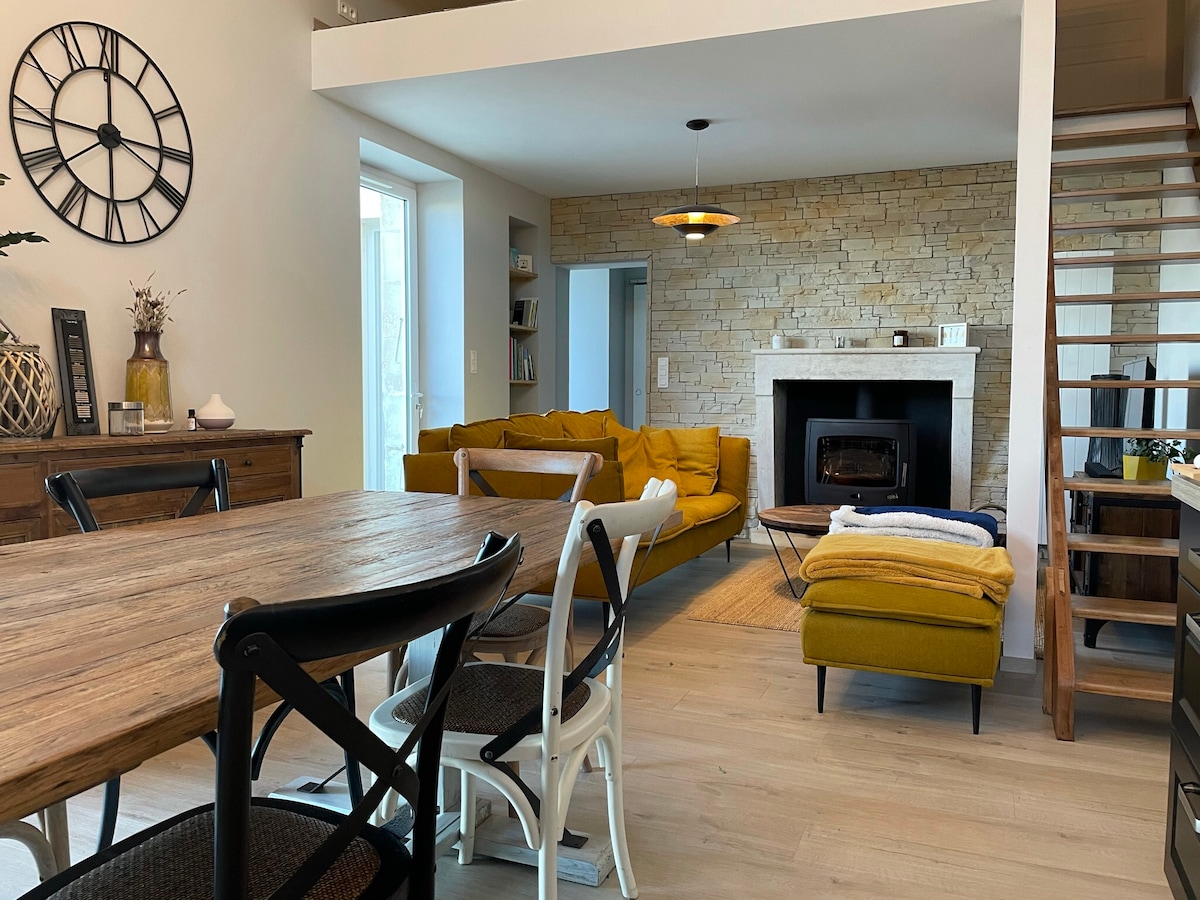 A bright living space features a yellow sofa and a warm fireplace, creating a cozy atmosphere. Light wooden floors lead to a dining area with a rustic table surrounded by diverse chairs. A staircase with wooden accents leads to the upper level.