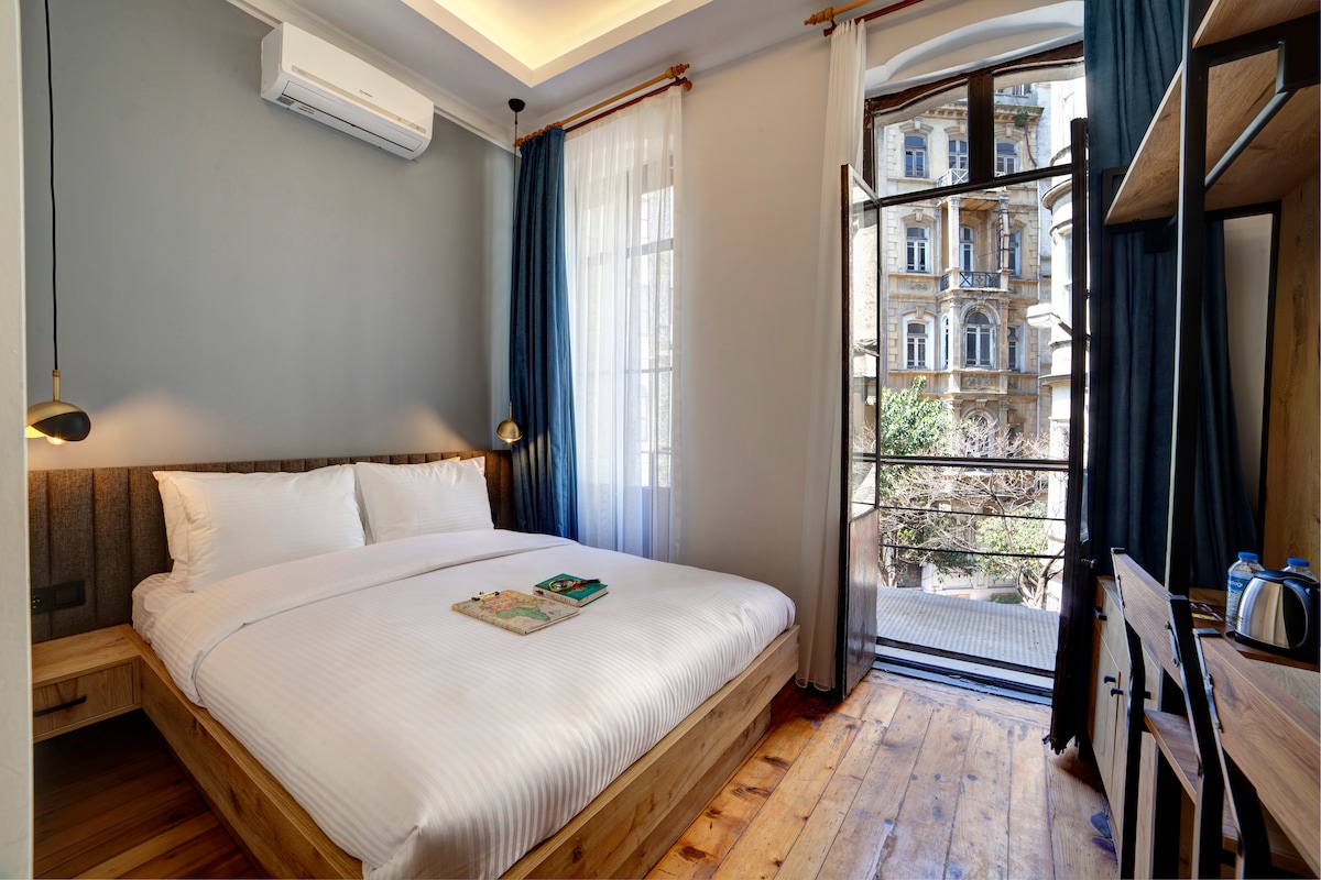 A well-lit room features a wooden bed with a neatly arranged white comforter. Large windows allow natural light to fill the space, offering views of the outdoor greenery. A small desk and chair are positioned against the wall, creating a functional workspace.