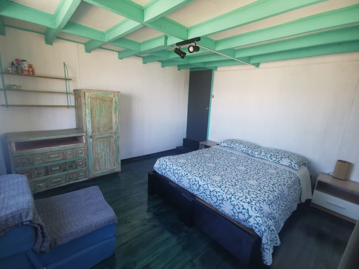 A stylish room features a spacious bed dressed in patterned linens. A vintage dresser stands against one wall, while a cozy seating area is positioned nearby. Light streams in, highlighting the turquoise-painted ceiling and polished floor, creating a serene environment.