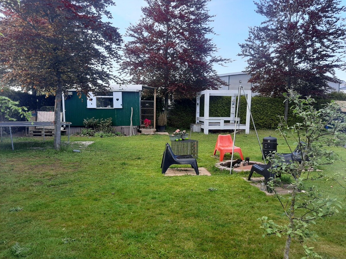 A green garden space is presented with a variety of trees and plants. Several lounge chairs are arranged around a fire pit, while a swing set and a small table are visible. The pipowagen and a seating area can be seen in the background.