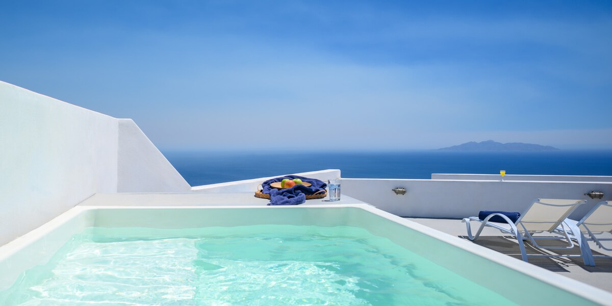 A private terrace is highlighted by a hot tub surrounded by clear blue water. Sun loungers are positioned nearby, with a platter of colorful fruits presented on a towel. The expansive view of the ocean and clear blue sky creates a serene ambiance.