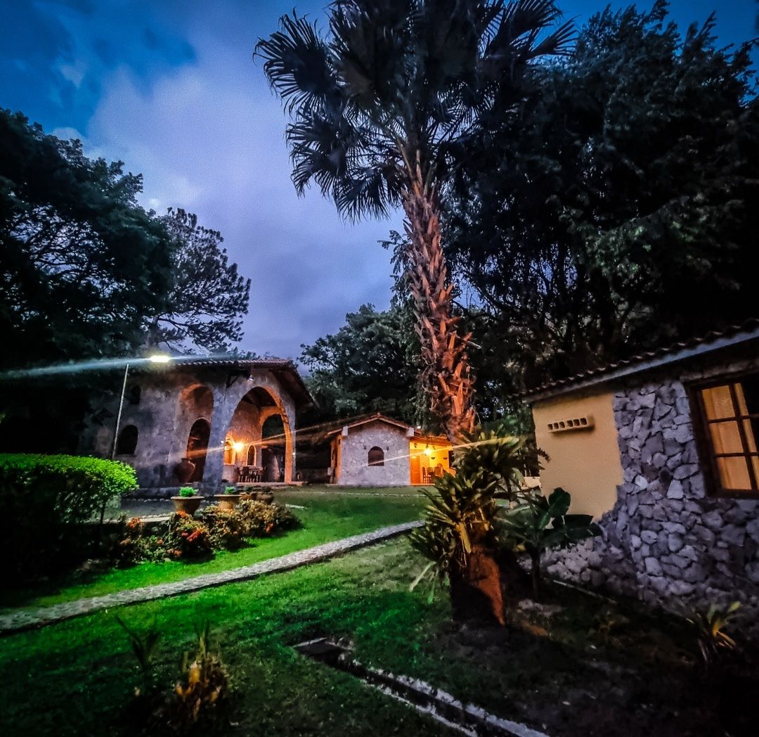 A serene outdoor setting features a stone building with arched openings, surrounded by lush greenery and illuminated by soft outdoor lighting. A palm tree stands tall, adding to the tranquil ambiance. Well-maintained pathways and plants enhance the inviting atmosphere of the property.
