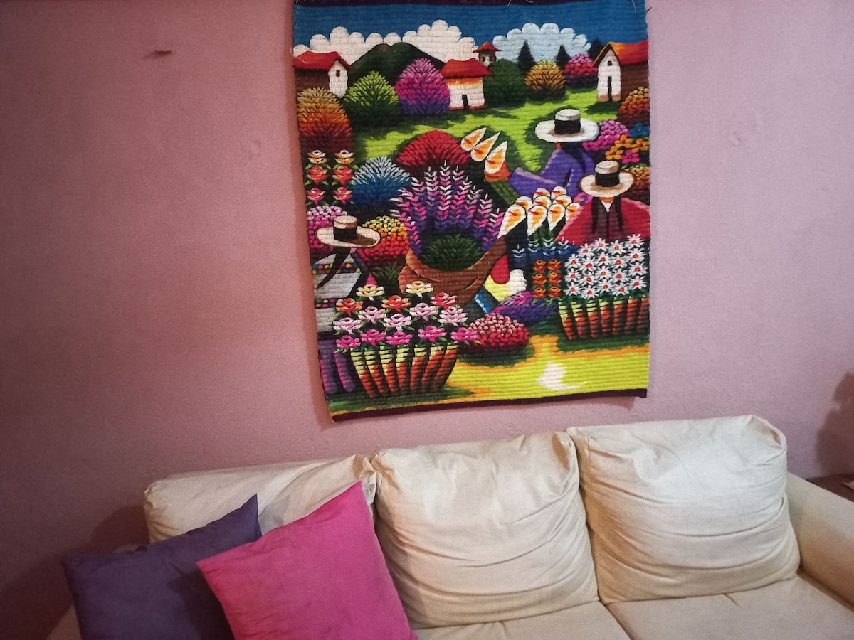 A colorful wall tapestry depicting a vibrant market scene is showcased above a light-colored sofa. The artwork features lush plants, flowers, and figures in traditional attire, adding a local cultural touch to the space. Decorative cushions in shades of purple are arranged on the sofa.