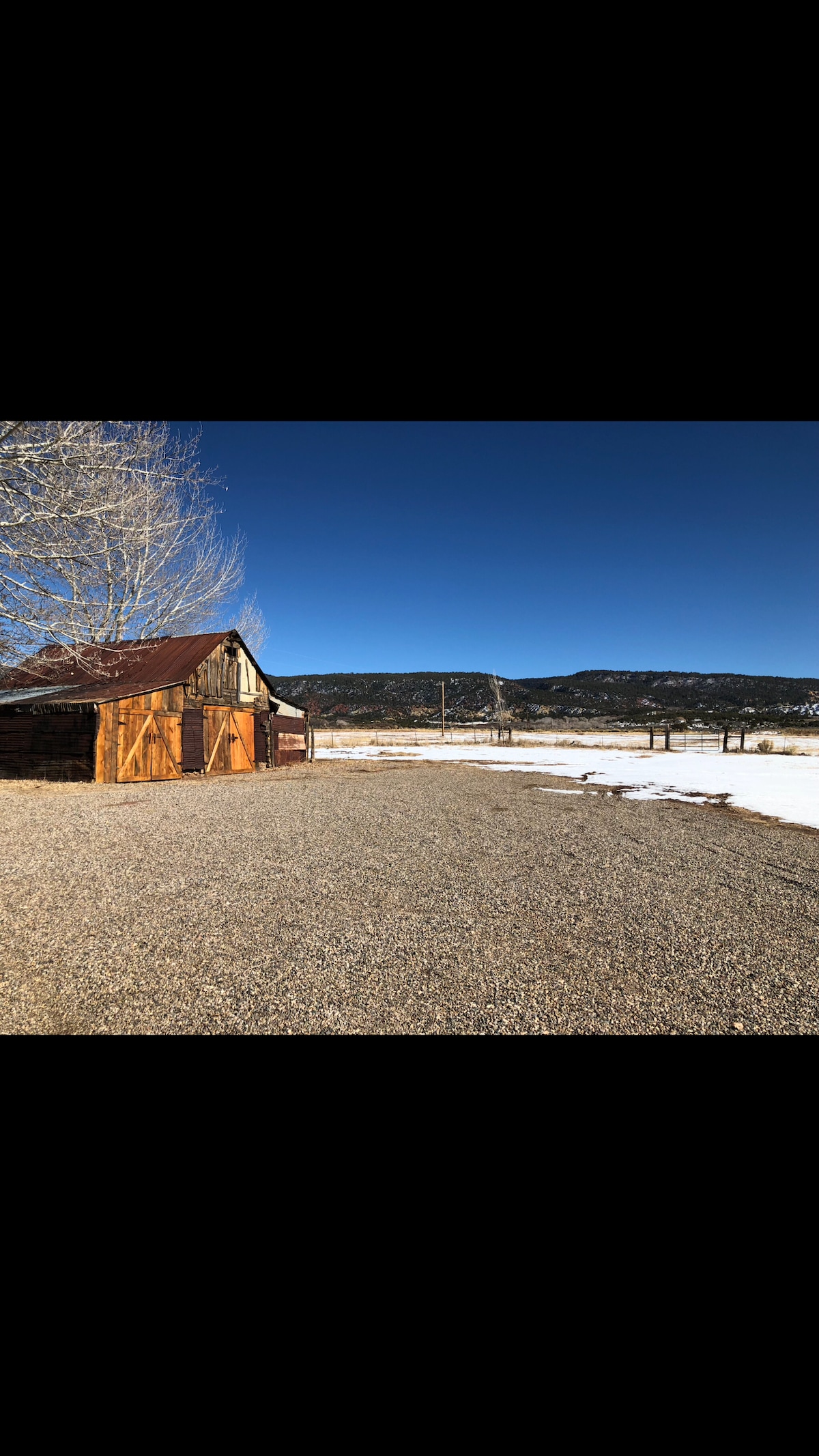 A rustic building stands on a gravel surface, surrounded by an open landscape. The clear blue sky contrasts with the mountainous backdrop, and hints of snowy ground are visible, suggesting a serene and tranquil environment.