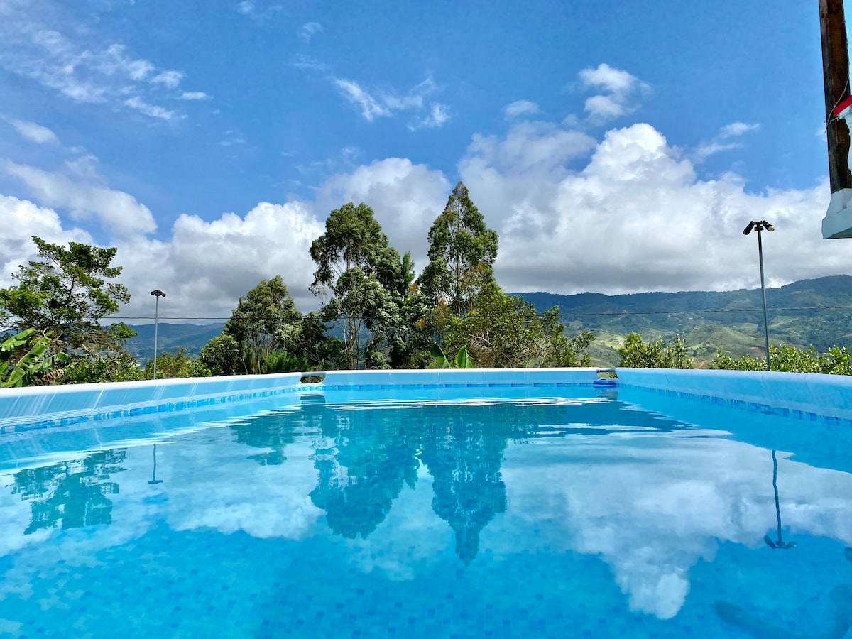 A refreshing swimming pool reflects the blue sky and surrounding greenery, framed by tall trees and distant mountains. Soft clouds float above, enhancing the natural beauty of the landscape.