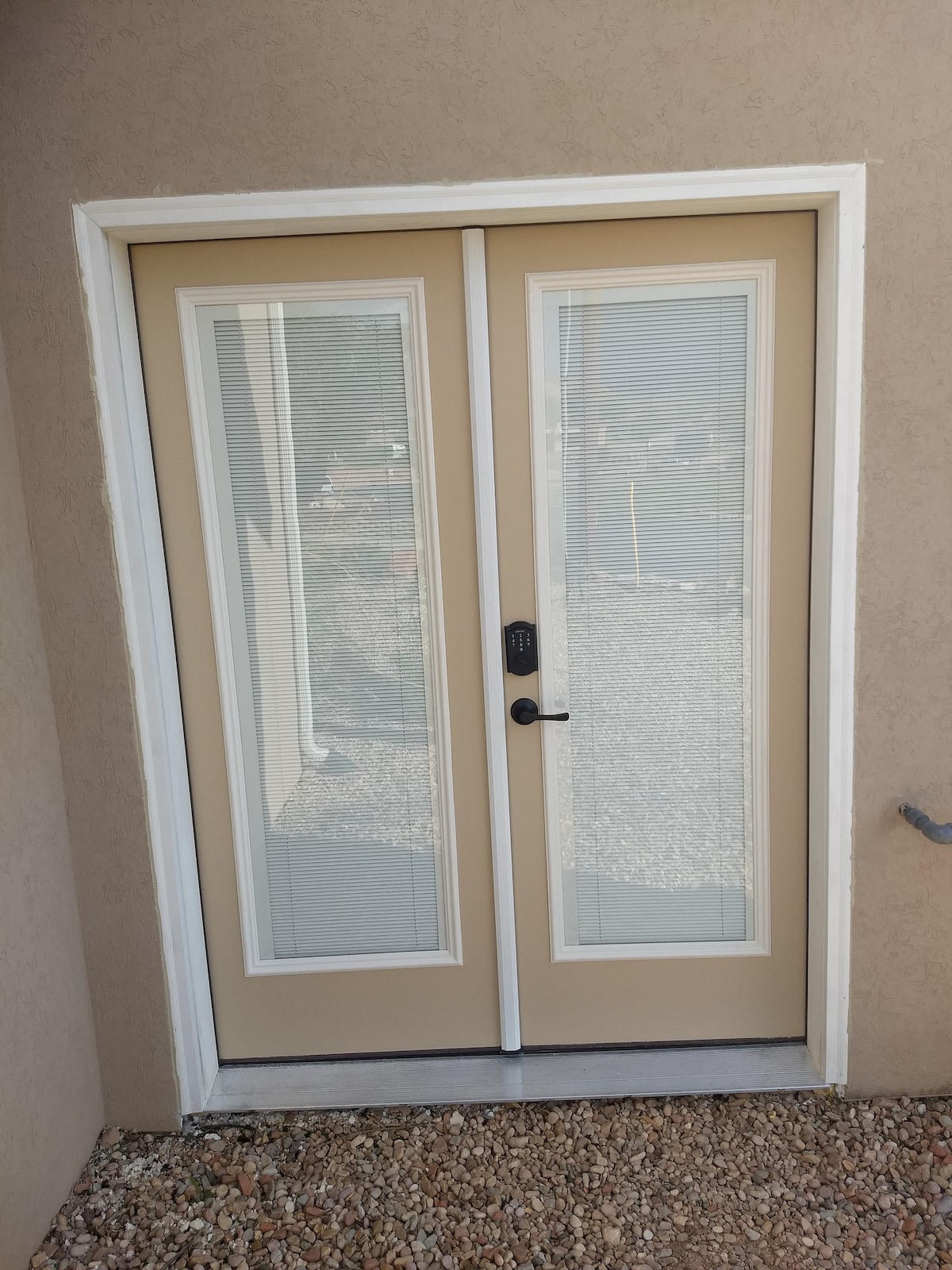 A set of double glass doors with a beige frame is visible, featuring horizontal blinds between the panes. The entrance leads to a private space, with a pebbled area surrounding the door, providing easy access to the outdoors.