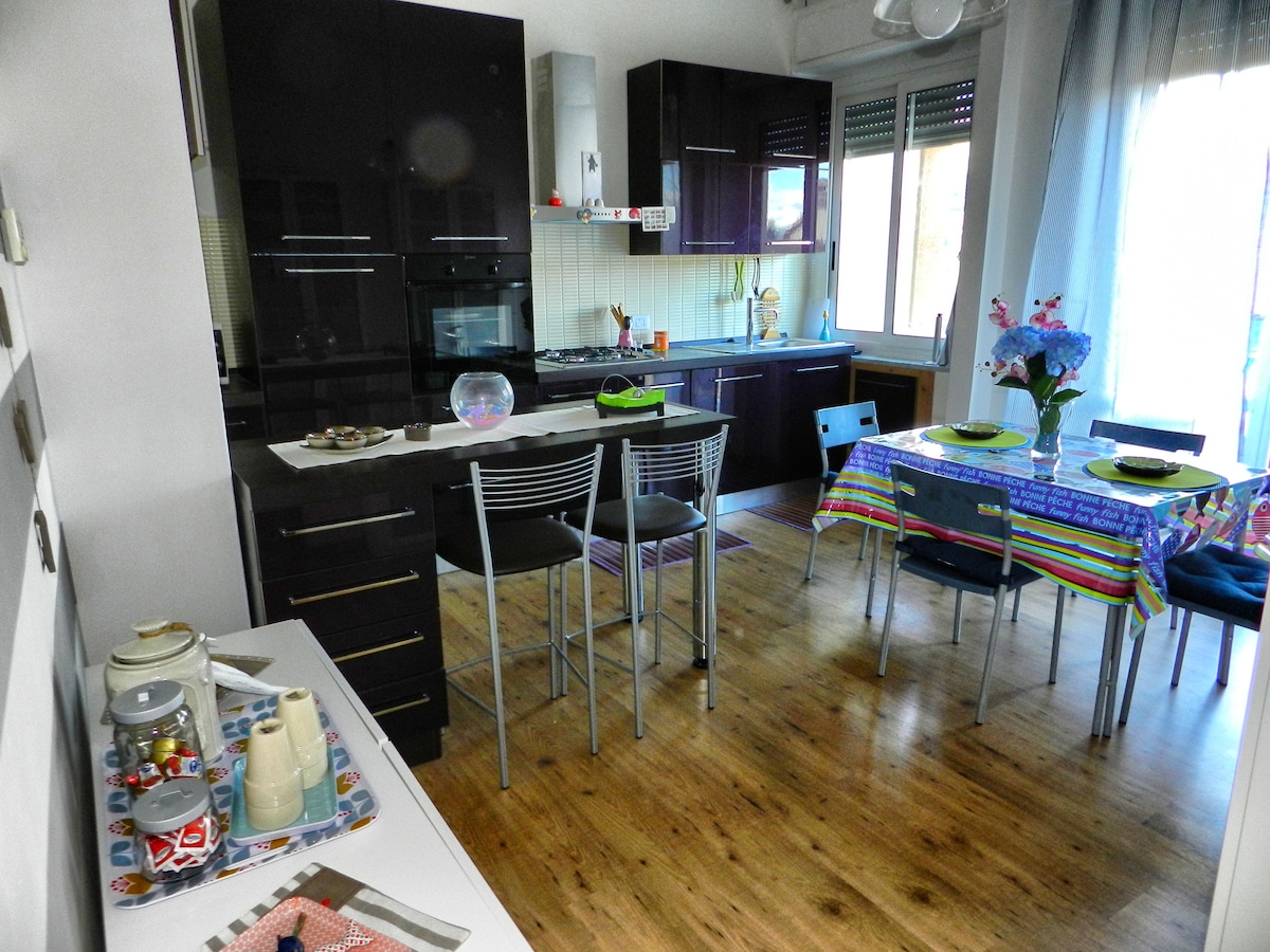 The kitchen area features dark cabinetry and modern appliances, including an oven and a refrigerator. A dining table is set with colorful plates, while bar stools are positioned at the counter. Natural light streams in through the window, enhancing the warm wood flooring.