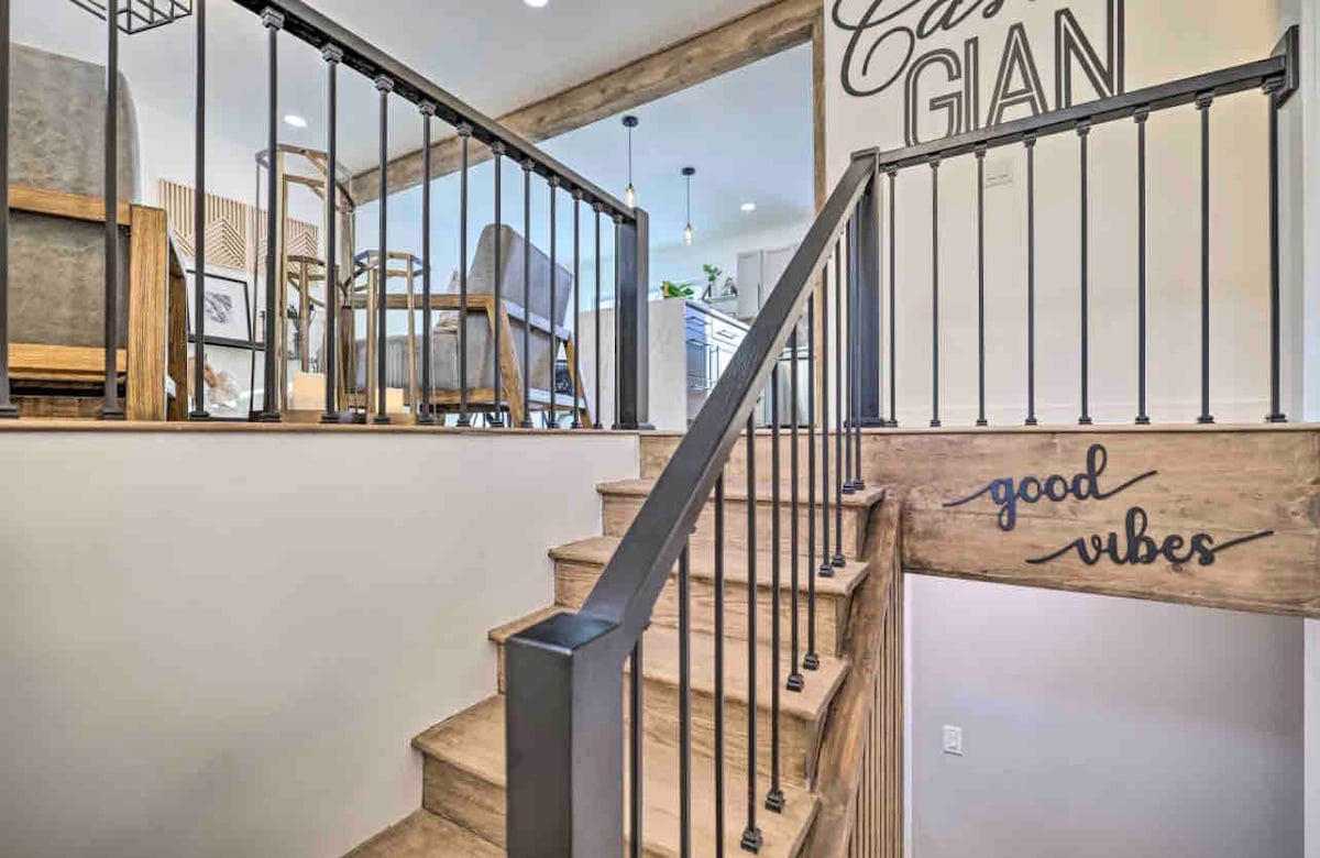 A wooden staircase with black metal railings leads to a light-filled open living area. The wall displays a motivational phrase, while the surrounding space features modern furniture and greenery, creating an inviting transition between levels.