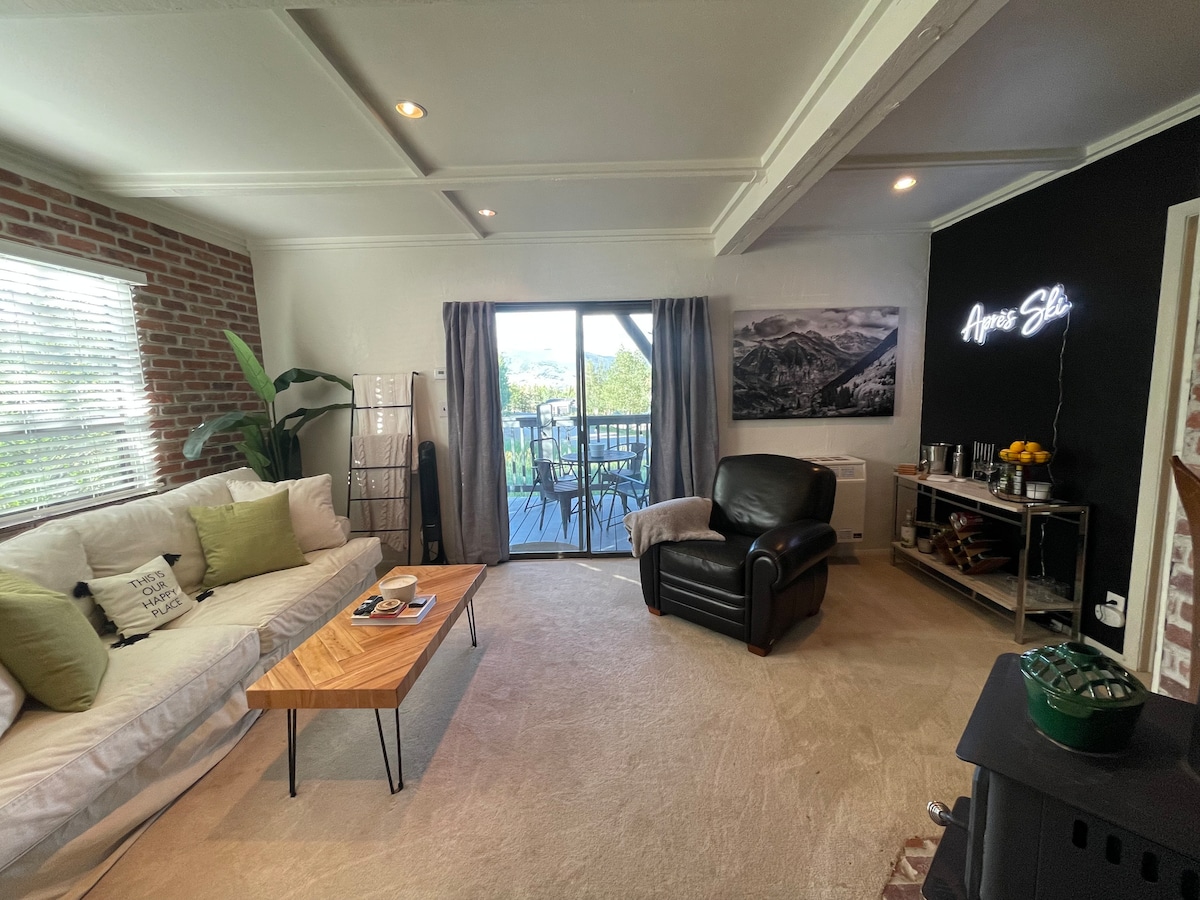 A comfortable living area is presented with a light-colored sofa and a stylish wooden coffee table. Large glass doors offer a view of the outdoor space, while a black accent wall is adorned with a mountain-themed artwork. A cozy leather chair complements the space.