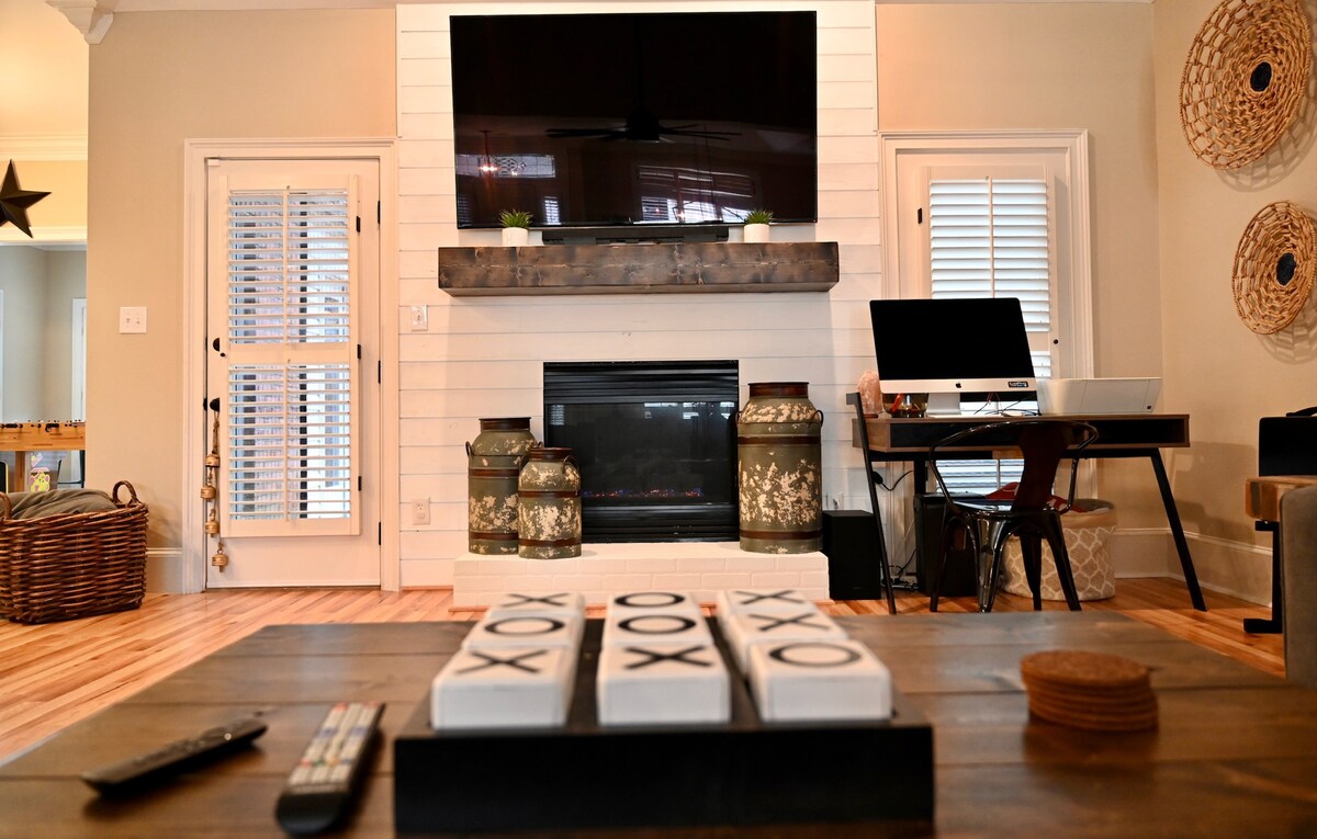 A modern living space features a mounted flat-screen TV above a sleek fireplace. A wooden shelf displays decorative items, while a coffee table with a game set is positioned in the foreground. A desk and chair are visible in the background, enhancing the room's multifunctional appeal.