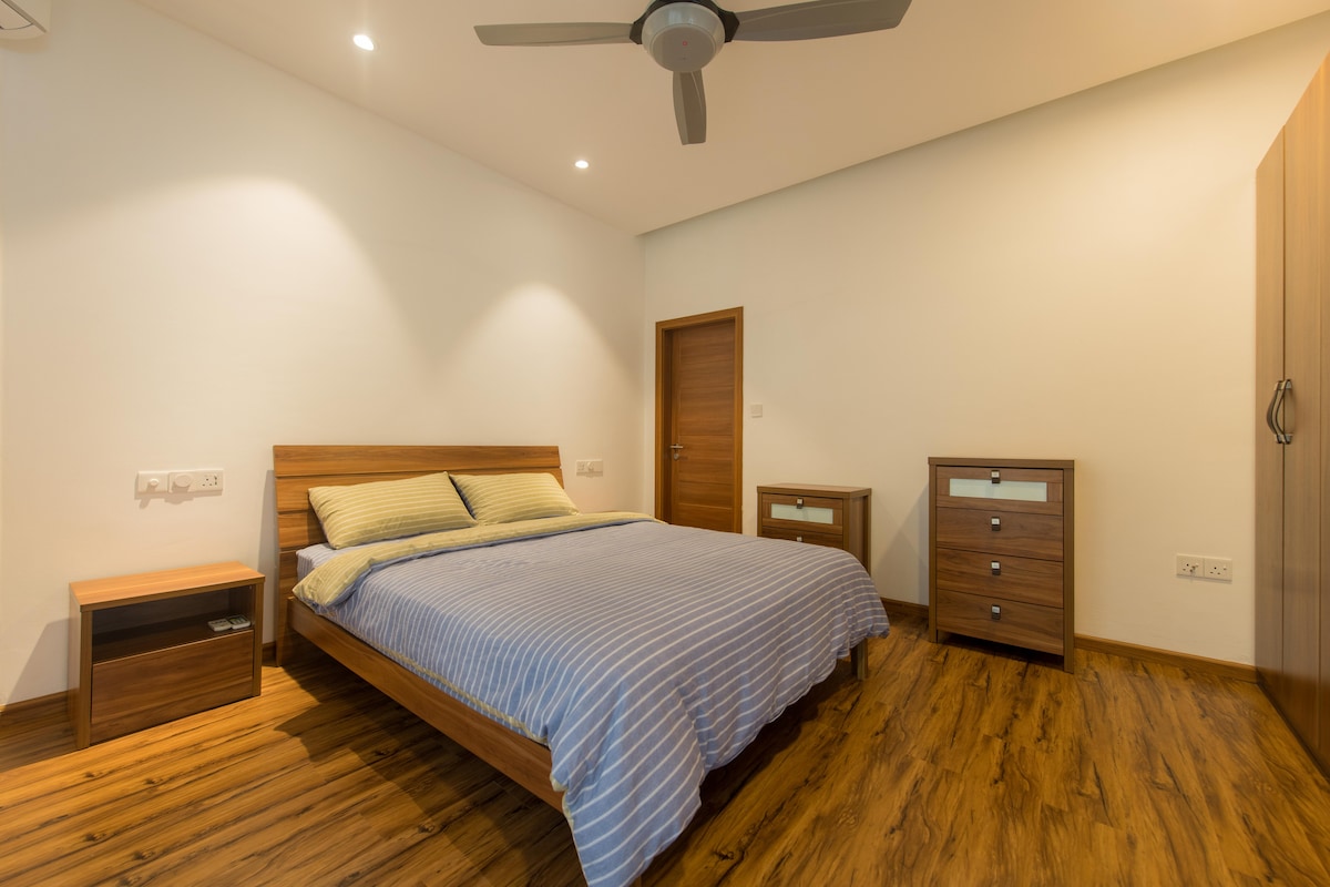 A spacious bedroom is presented with a wooden bed frame and a striped blanket. A bedside table and a chest of drawers complement the room, while warm wood flooring adds a natural touch. Soft ceiling lights provide illumination, and a ceiling fan enhances airflow.