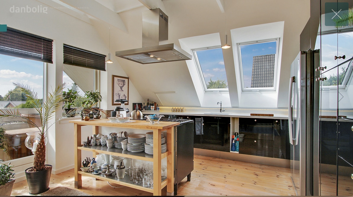 A modern kitchen area features an island with wooden countertops, displaying various dishware and glassware. The space is brightened by large skylights, allowing natural light to fill the sleek, dark cabinets. A potted plant adds a touch of greenery to the environment.