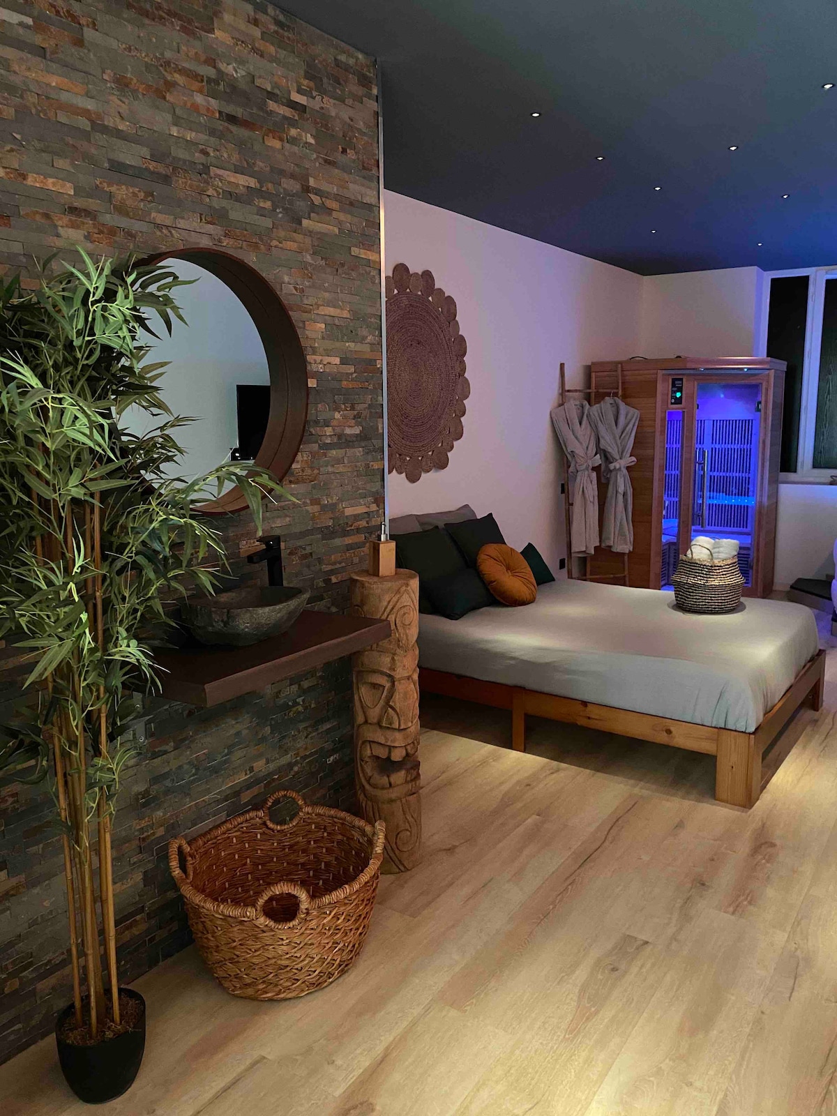A cozy room is presented with a wooden bed, complemented by natural tones and stone accent walls. A decorative mirror reflects soft lighting, while a sauna and relaxation area are visible in the background. A woven basket adds a touch of texture to the space.