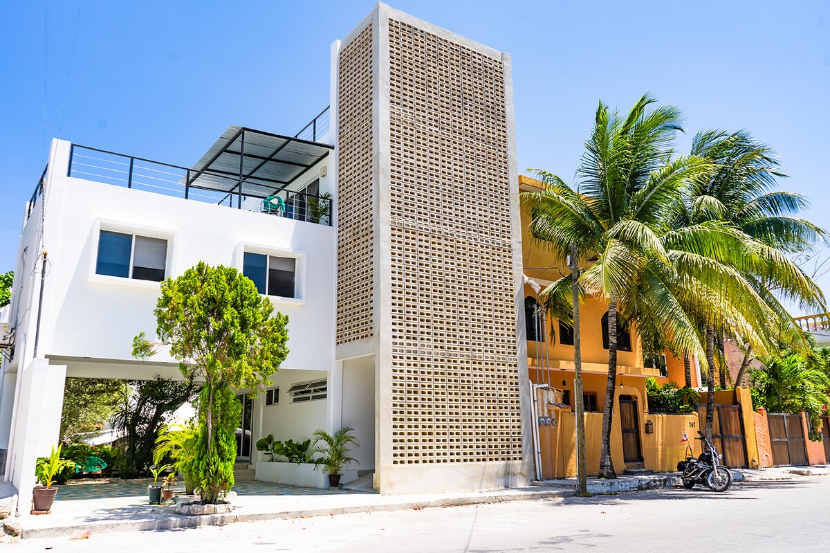 A modern building rises with a contemporary facade, featuring a distinctive geometric design. Lush greenery surrounds the entrance, while large windows provide natural light. A balcony is visible on the upper level, offering an outdoor space. Palm trees line the street, enhancing the tropical atmosphere.