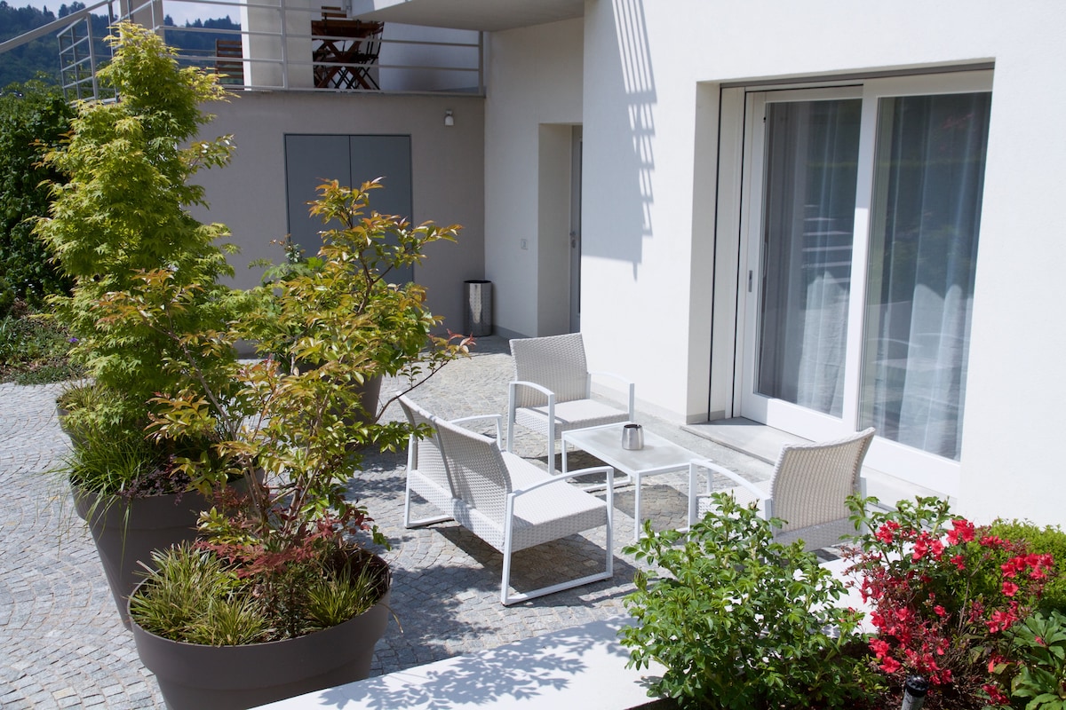 A private patio is surrounded by vibrant plants and greenery. The area features a small seating arrangement with white furniture, including a table and chairs. Large potted plants add layers of color, while sliding glass doors lead into the living space.