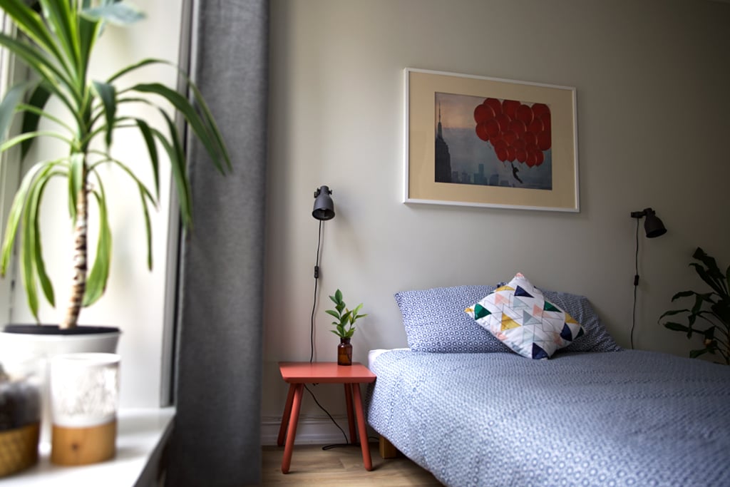A bright bedroom features a comfortable double bed adorned with a patterned pillow. A vibrant red side table holds a small potted plant. Light enters through a nearby window, while decorative art hangs above the bed, adding a touch of character to the space.