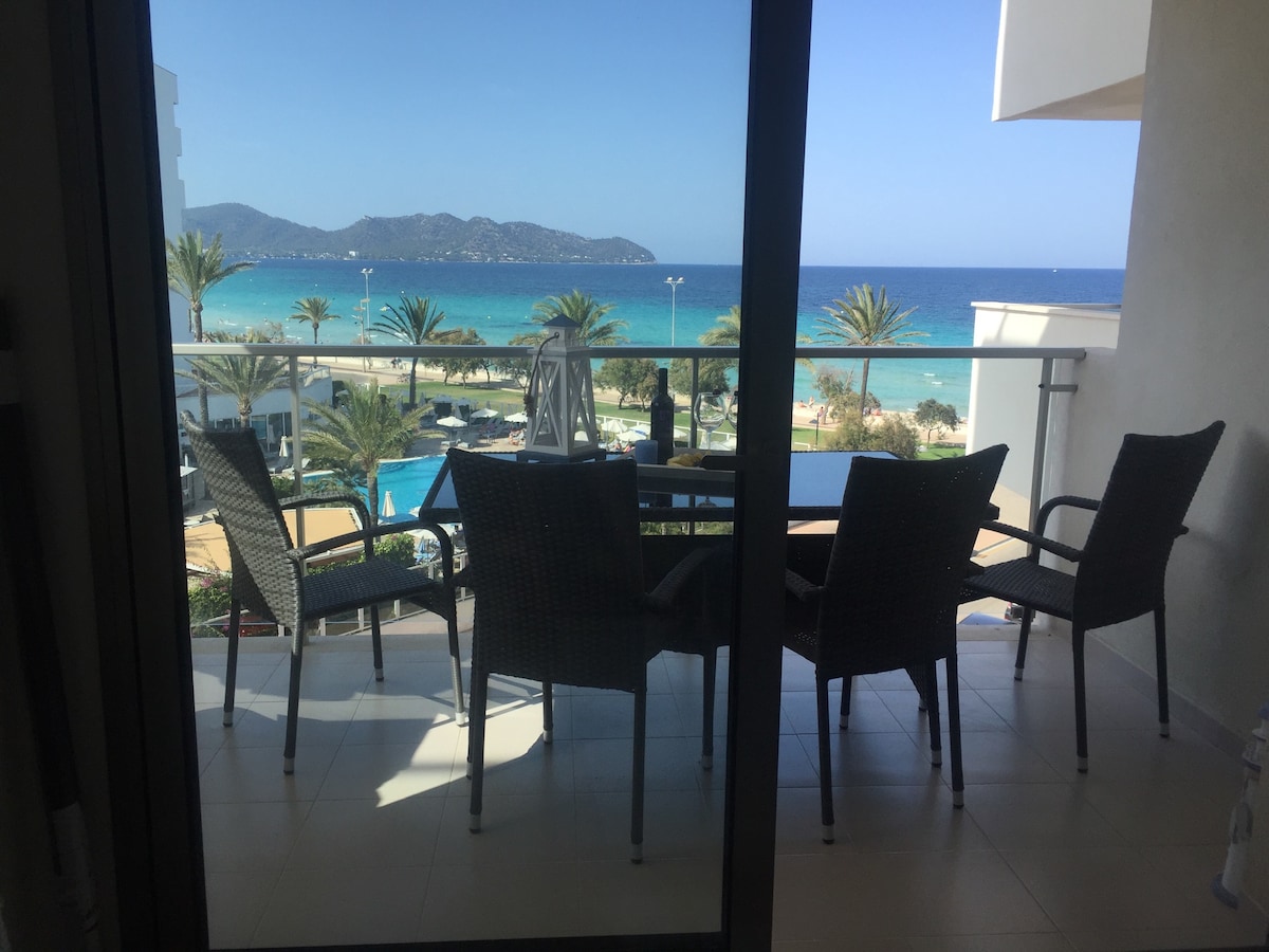 A terrace is observed through large glass doors, featuring a dining table with four woven chairs. A panoramic view of the Mediterranean Sea can be seen in the background, framed by palm trees and the coastline.