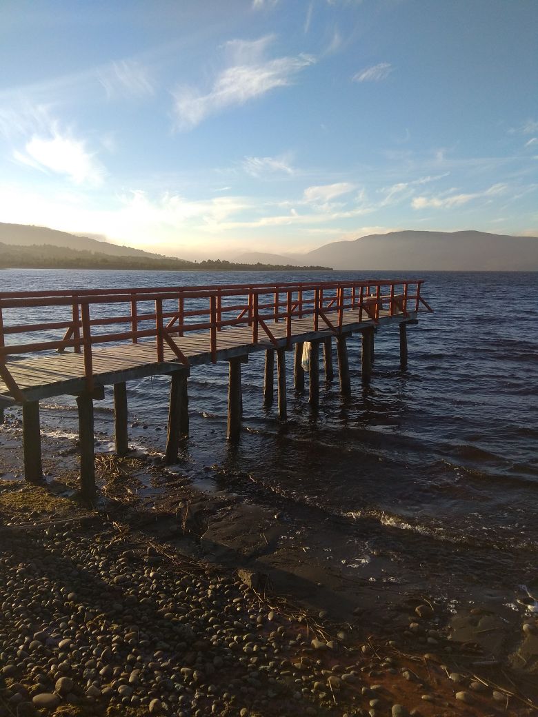 A wooden dock extends over the tranquil waters of Lake Huillinco, supported by sturdy posts. Sunlight casts a warm glow, illuminating the surrounding landscape of hills in the distance. Pebbles line the edge of the shoreline, adding a natural element to the serene setting.
