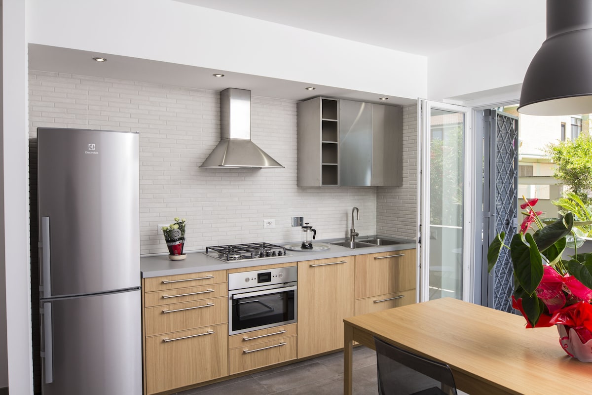 The modern kitchen features sleek cabinetry and stainless steel appliances, including a refrigerator and an oven. A gas stove with a streamlined hood is positioned above, while a dining table with chairs offers a welcoming space. Natural light is welcomed through the open doorway.