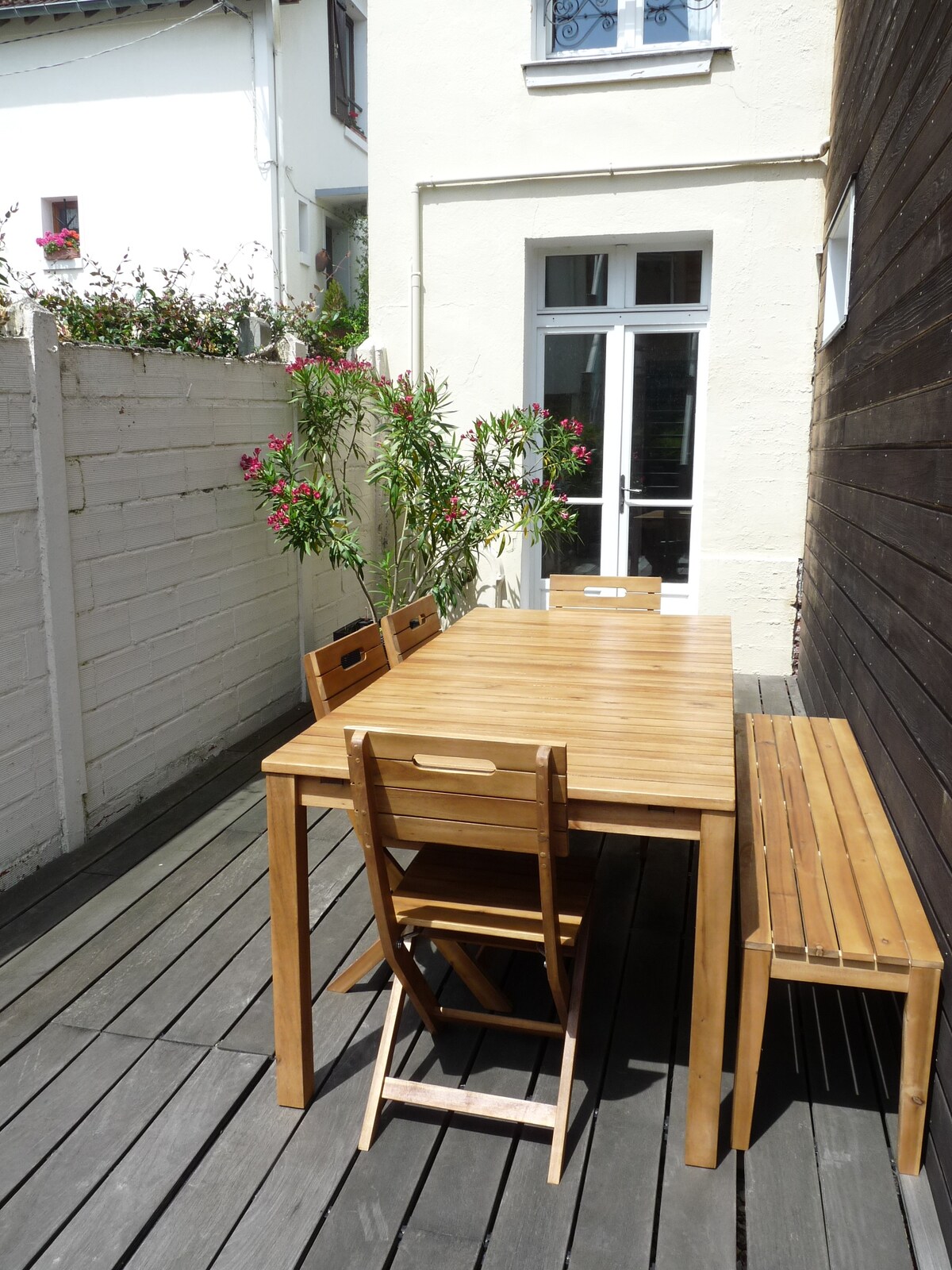 A wooden table with several seats is positioned on a deck area, surrounded by lush greenery. Bright flowers are visible near the walls, contributing to a serene outdoor space. Large windows can be seen in the background, allowing natural light to enter.