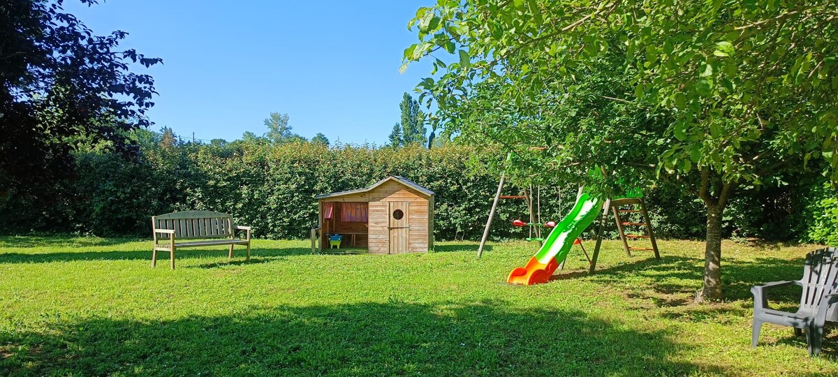 A spacious outdoor area is filled with green grass and bordered by dense hedges. A wooden playhouse is visible, accompanied by a colorful slide and a swing set. Seating options include a wooden bench and a white chair, offering relaxation amid the lush environment.