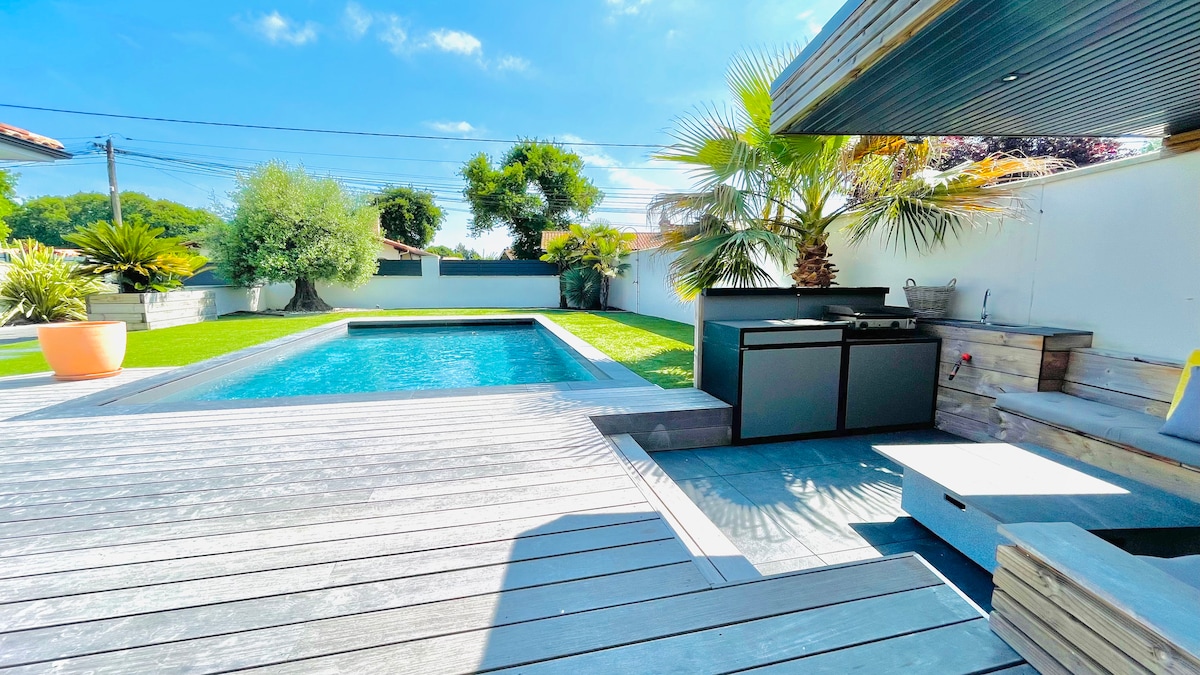 A serene outdoor space is highlighted, featuring a clear salt-water pool surrounded by greenery. A wooden deck complements the area, with a shaded lounge setup and an outdoor kitchen unit equipped with a barbecue, creating an ideal setting for relaxation.
