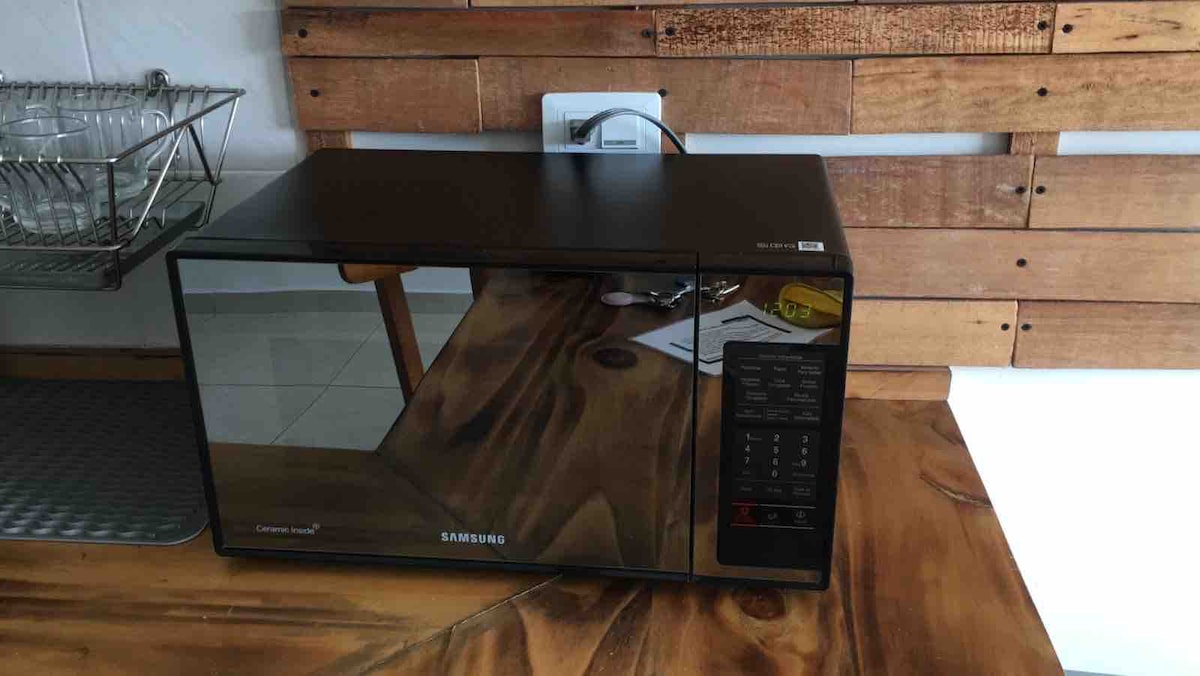 A sleek, black Samsung microwave is situated on a wooden countertop. The design features a glass door allowing visibility of the interior. Nearby, an organized dish rack is placed against a textured wooden wall, contributing to a functional kitchen environment.