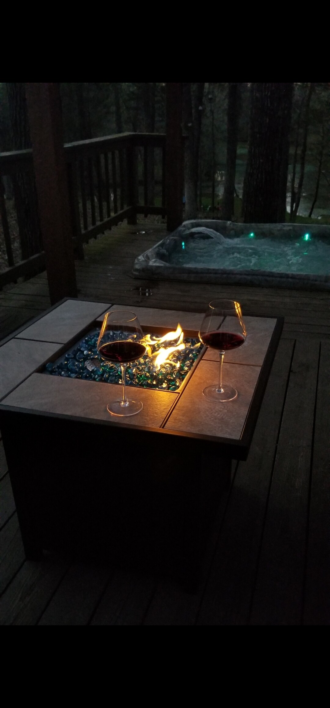 A cozy outdoor setting is captured with a fire pit glowing softly atop a square table, surrounded by glass stones. Two glasses of red wine are placed on the table, adding a touch of elegance. A hot tub is visible in the background, subtly illuminated.