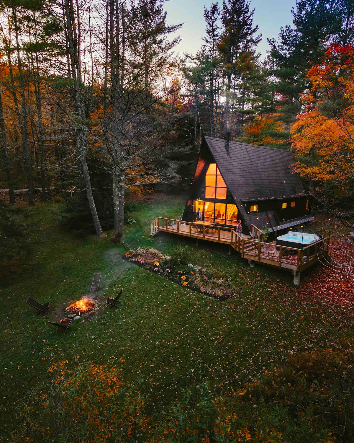 The A-frame cabin is nestled among vibrant autumn foliage, with large windows reflecting warm light. A hot tub is positioned on the deck, which overlooks a well-kept lawn. A fire pit area surrounded by seating is situated near the cabin.