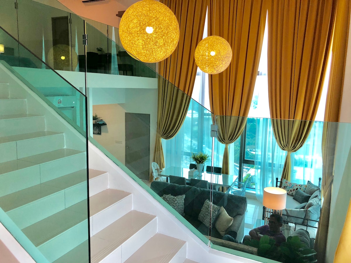 A modern staircase features sleek white steps, accompanied by a glass railing. Bright yellow curtains frame large windows, allowing natural light to illuminate the living area below, which contains a comfortable seating arrangement and decorative plants.