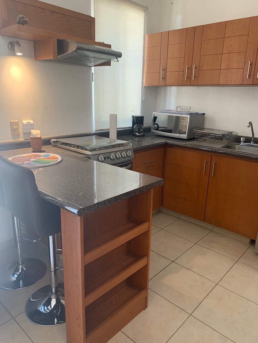 A modern kitchen features wooden cabinets and a granite countertop. Stainless steel appliances, including a stove and microwave, are visible. A small breakfast bar with two sleek stools offers a casual dining option. Natural light filters in through a nearby window.