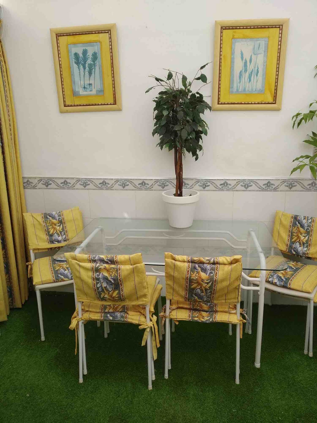 A small dining area is adorned with a glass table surrounded by four yellow and blue patterned chairs. A potted plant occupies the center of the table, while framed artwork hangs on the wall, adding a touch of color to the room.