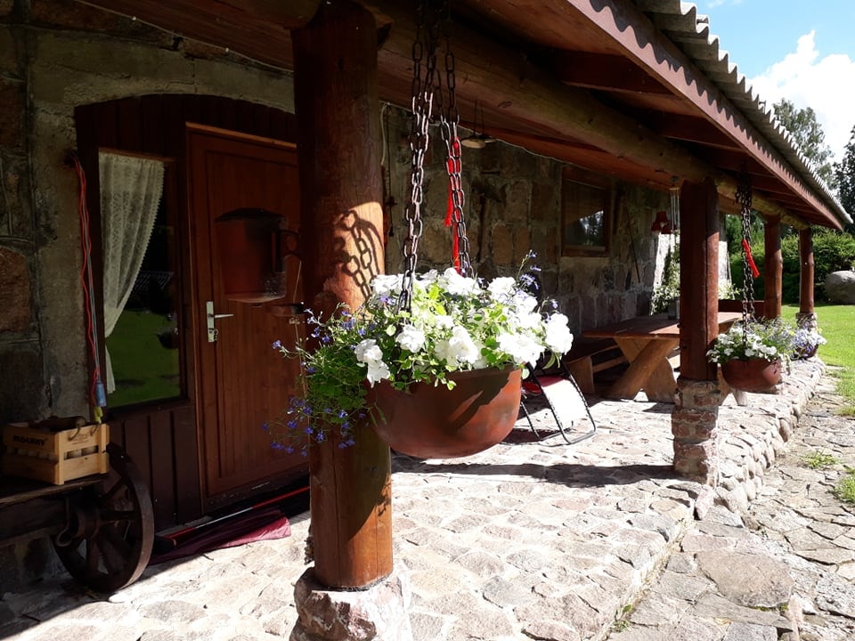 The entrance is adorned with hanging flower pots filled with white and purple blooms. Natural stone and wooden beams frame the doorway, which opens to a cozy interior. A rustic wooden bench is positioned nearby on the stone pathway.