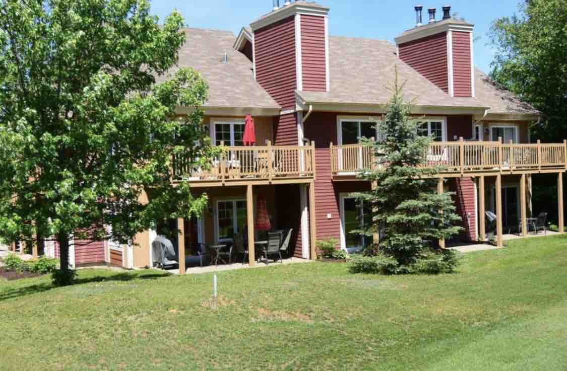 The exterior of a two-story condo features rich red siding and multiple balconies. An inviting wooden deck can be seen with outdoor seating arrangements. Lush greenery surrounds the property, complemented by tall trees and manicured lawns, creating a peaceful outdoor environment.