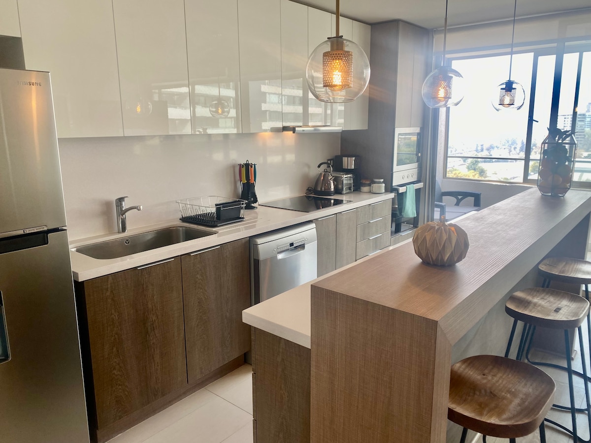 A modern kitchen is showcased with sleek cabinetry and a spacious countertop. Stainless steel appliances, including a dishwasher, are visible, along with colorful kitchen utensils. A bar with wooden stools offers additional seating, while large windows provide natural light and views of the city.