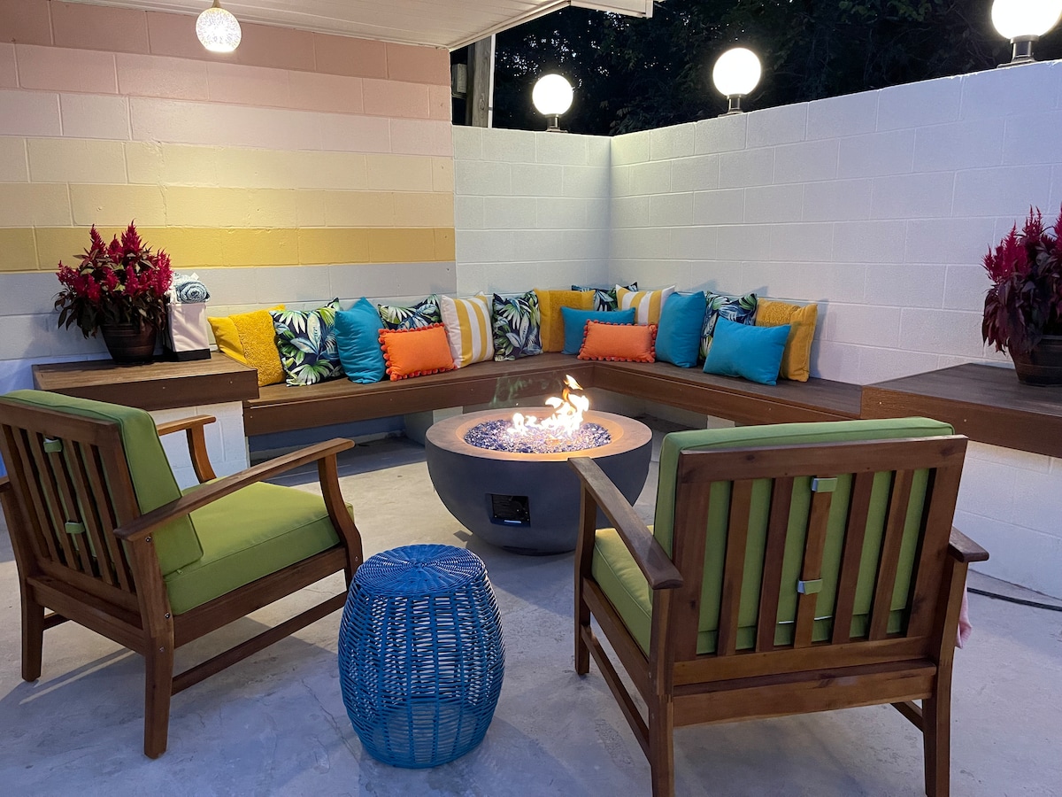 An inviting outdoor seating area features a circular fire pit surrounded by a cozy bench adorned with colorful cushions. Two wooden chairs with green cushions are positioned nearby, complemented by a blue accent table. The surrounding walls are decorated with soft pastel colors and potted plants.