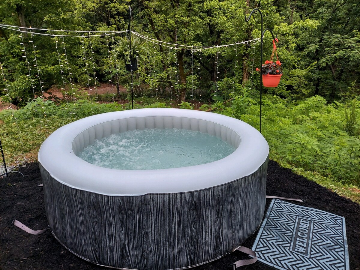 An inflatable hot tub is positioned on a dark gravel surface, surrounded by greenery. String lights are visible overhead, and a hanging planter with flowers adds a touch of color. A welcome mat lies at the entrance, inviting relaxation in nature.
