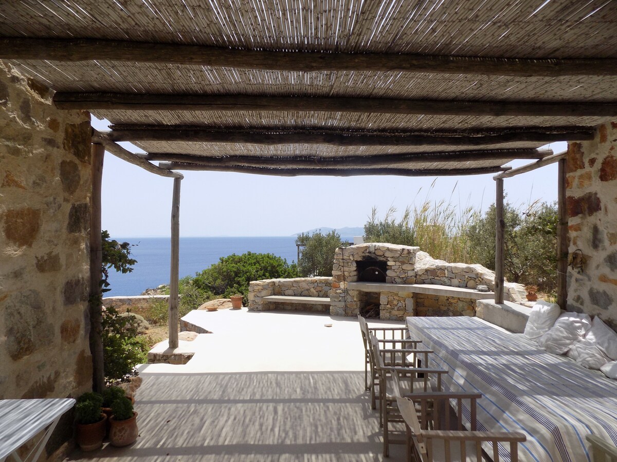 An outdoor seating area covered by a thatched roof provides a view of the sea. A stone oven is visible in the background, surrounded by greenery. Simple wooden furniture with light-colored cushions is arranged to create a relaxed environment for gatherings.