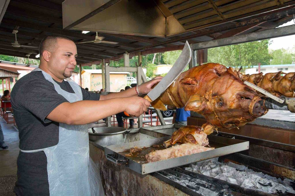 A chef is seen skillfully carving a whole roasted hog at a well-equipped outdoor grill. The scene is enhanced by a setting that includes a covered workspace and a backdrop filled with greenery. Various cooking equipment is visible, indicating a busy culinary environment.