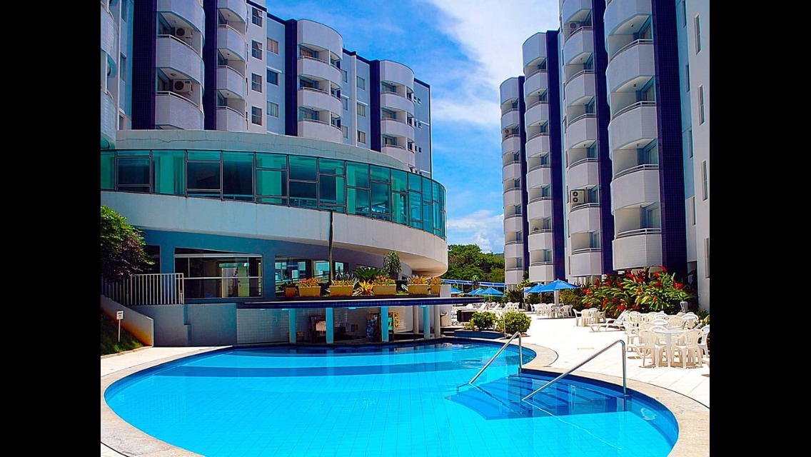 The image displays a spacious swimming pool surrounded by a well-kept area featuring lounge chairs. The modern buildings, with balconies, are visible in the background, complemented by tropical landscaping. The scene showcases bright blue skies, creating a welcoming atmosphere for relaxation.