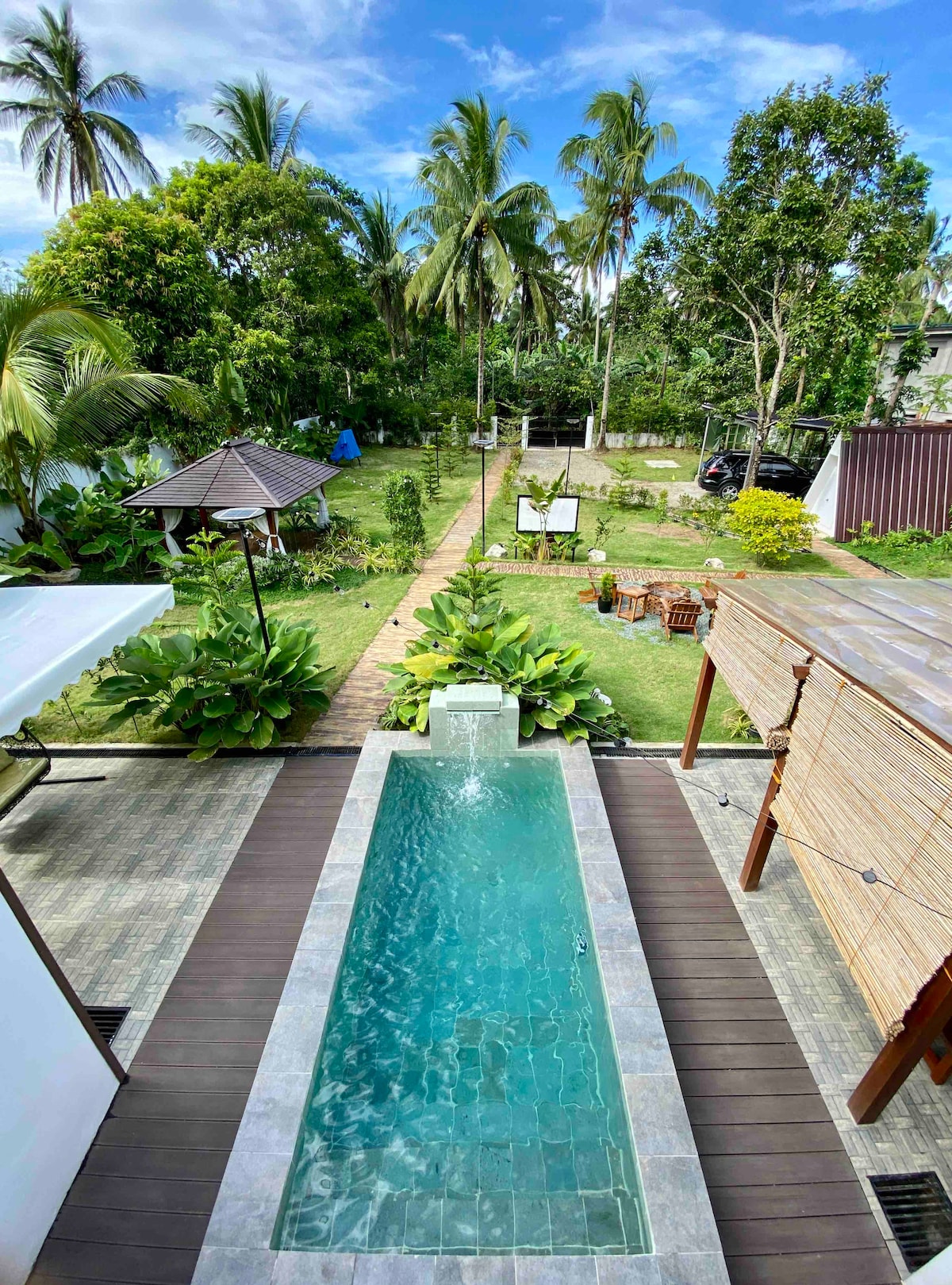 An inviting outdoor area features a narrow dipping pool bordered by greenery and a pathway leading through a lush garden. Palm trees and various plants provide natural shade and a serene environment. Relaxation spaces and shaded structures are visible in the distance.
