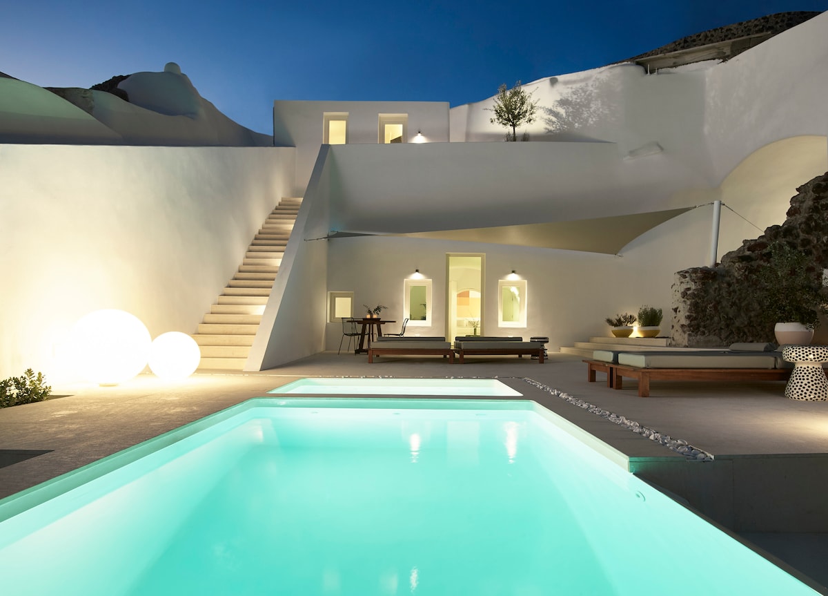 The outdoor area of the villa showcases two illuminated infinity pools, reflecting the serene blue of the water. Soft lighting enhances the whitewashed architecture, while stylish sun loungers surround the pool. A staircase leads to an upper level with additional living space, creating an inviting atmosphere.