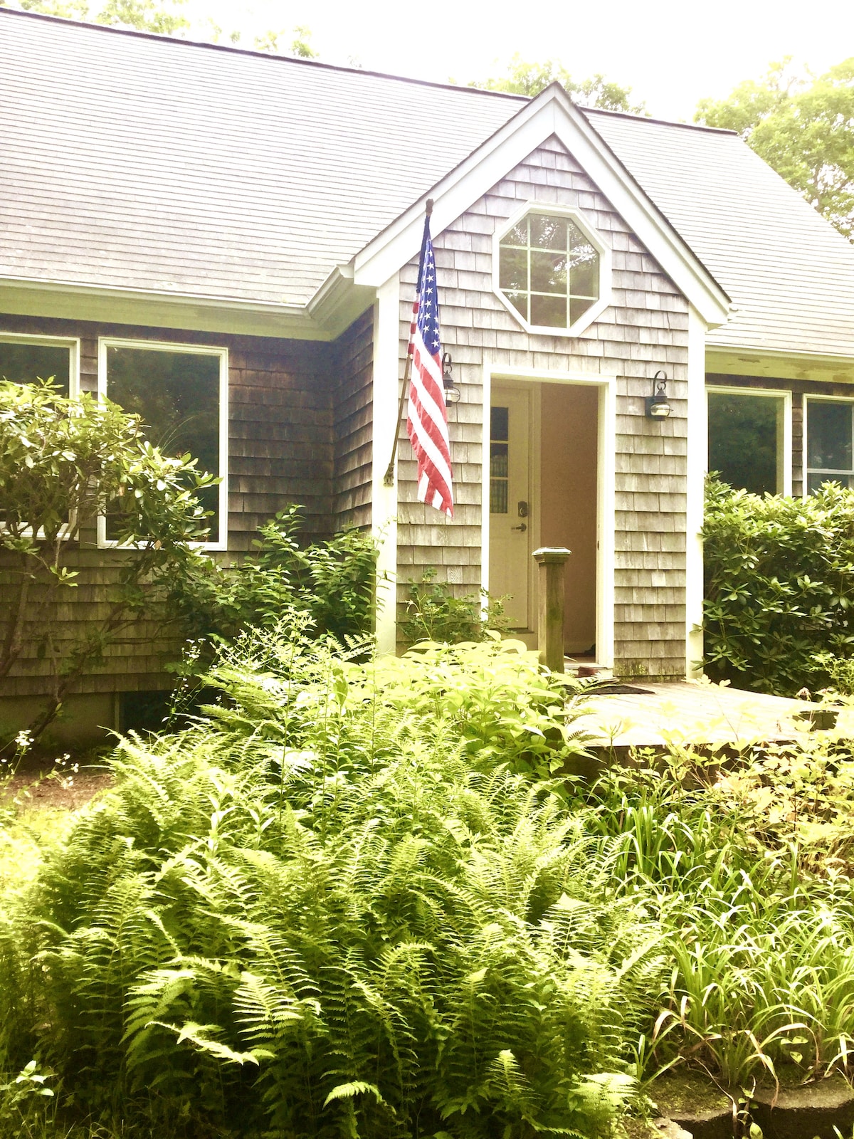 The entrance of the guest house is framed by lush greenery, featuring ferns and shrubs. A wooden path leads to a door with a round window above it. An American flag is prominently displayed, signaling a welcoming entry.