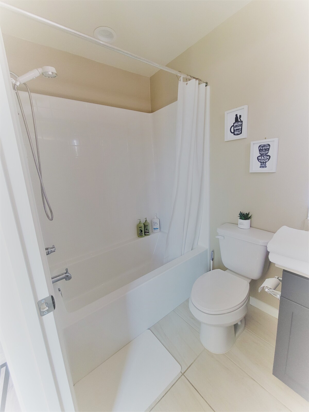 A clean and functional bathroom is depicted, featuring a bathtub with a shower curtain and a modern toilet. Personal care items are neatly arranged on the tub's edge. Subtle decor accents are visible on the wall, complementing the light-colored tiles.