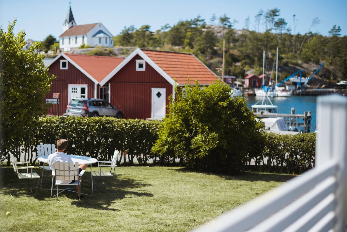 A peaceful lawn area is depicted with a white table and chairs overlooking the harbor. Red wooden boathouses and a few boats are visible in the background, surrounded by lush greenery and a distant church steeple. The scene captures a serene waterfront atmosphere.