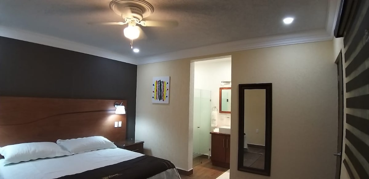 A well-appointed room features a spacious bed with white linens against a dark accent wall. A small nightstand is positioned beside the bed, equipped with a reading lamp. A bathroom area is visible in the background, with modern fixtures and glass walls enhancing the space.