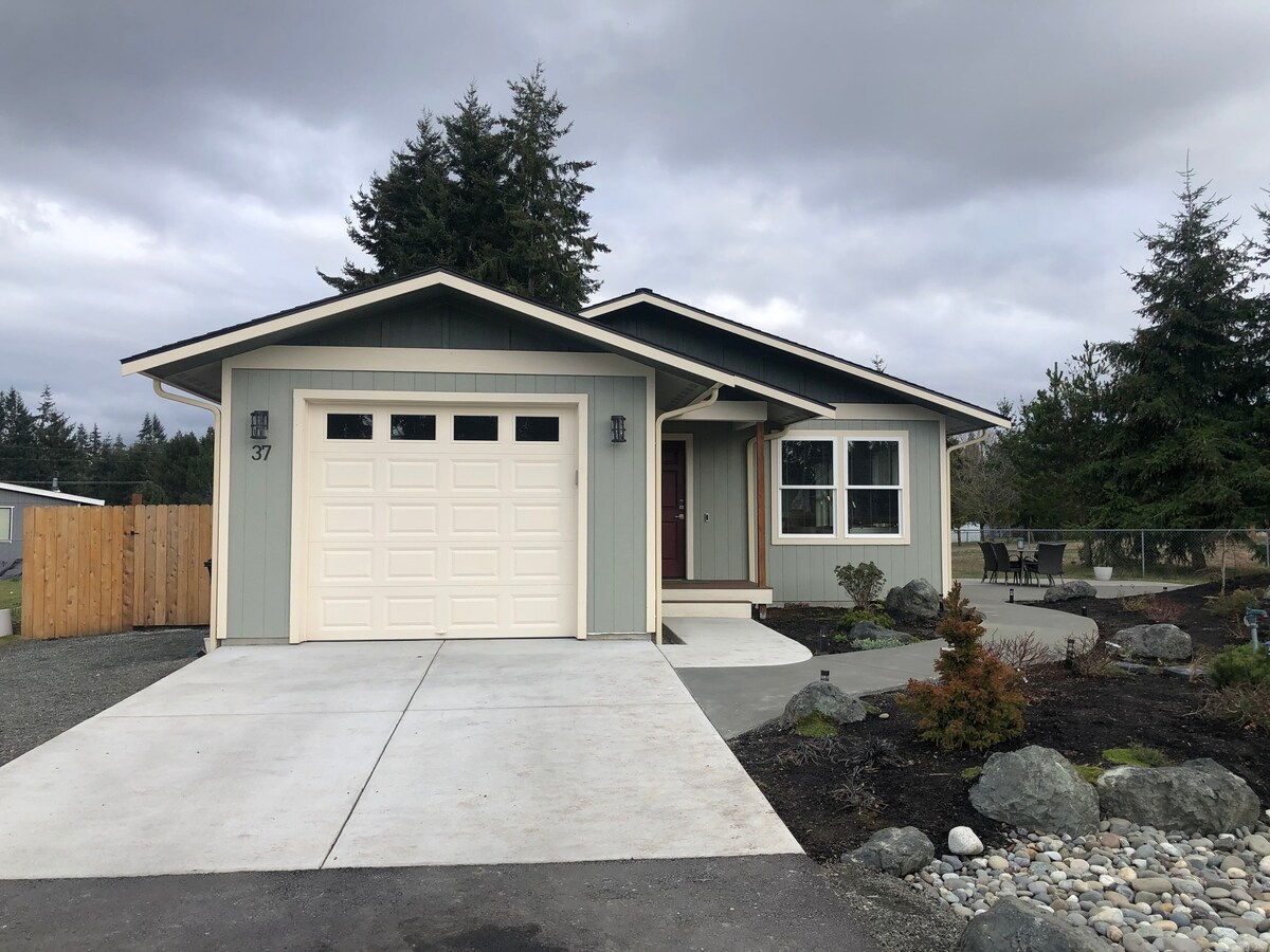 The exterior of the house presents a contemporary design with a light green facade and a white garage door. A paved driveway leads to the entrance, flanked by landscaped stones and small shrubs. Tall trees provide natural greenery in the background.