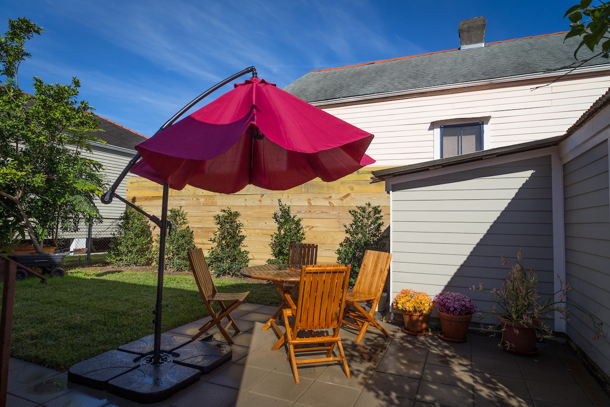 An outdoor patio area is equipped with a large, pink umbrella providing shade over a circular table surrounded by four wooden chairs. Potted plants and colorful flowers adorn the patio, with a lush green lawn in the background and wooden fencing offering privacy.