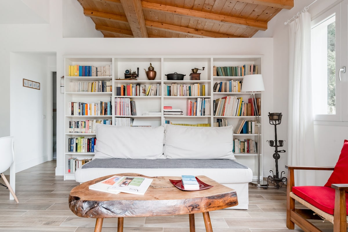 A bright living space features a white sofa with a gray accent cushion, positioned in front of a large bookshelf filled with a variety of books. A wooden coffee table sits nearby, complemented by a red upholstered chair and natural light streaming through a window.